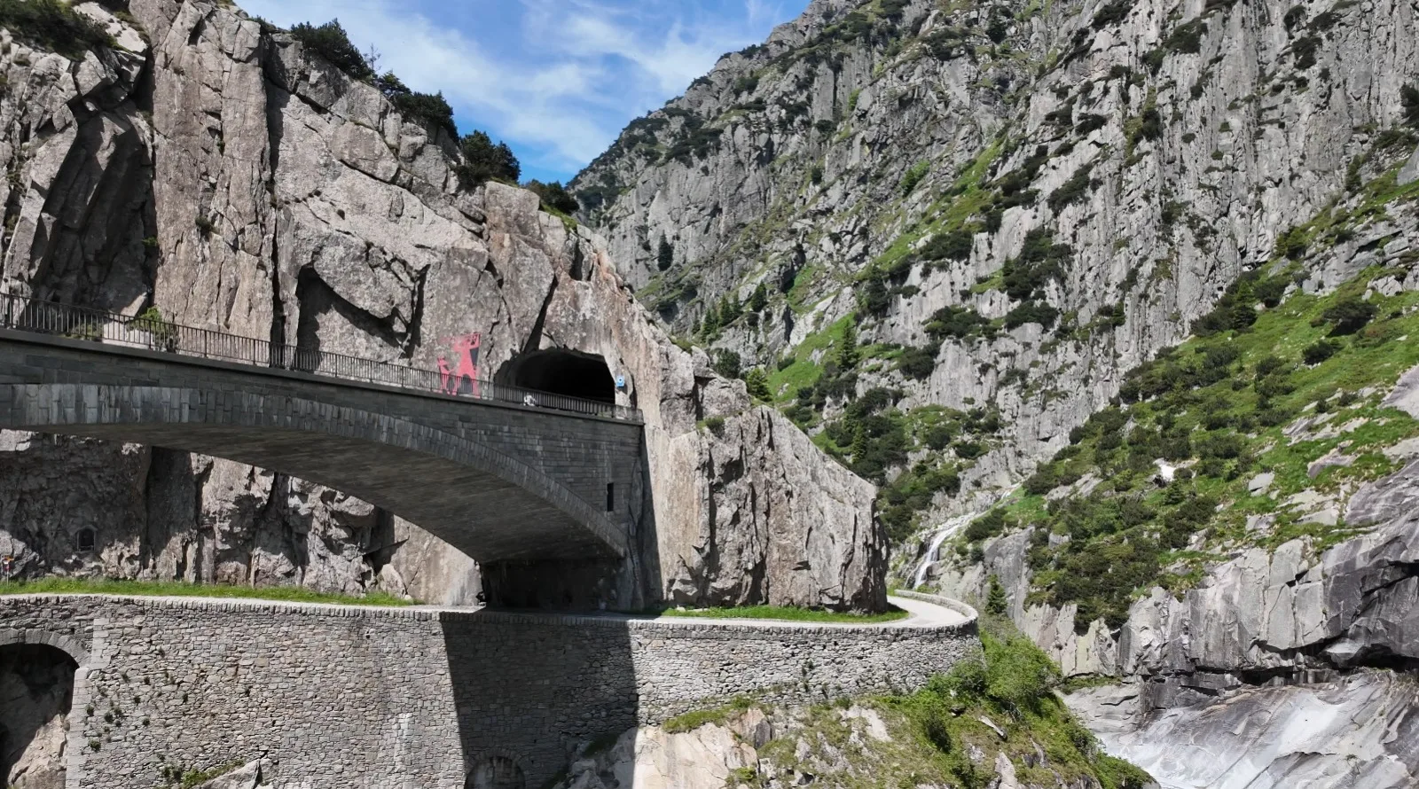 The Devil's Bridge in Andermatt — a stone arch bridge spanning the Schöllenen Gorge, with a pink devil silhouette mounted on the bridge wall and the Urnerloch tunnel mouth visible in the cliff to the right