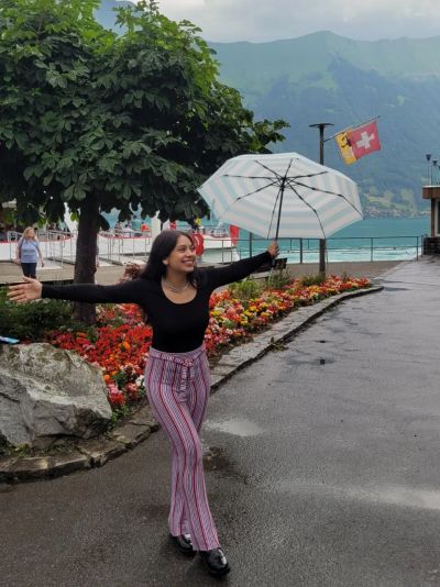 Traveler Jyoti holding a striped umbrella by Lake Brienz at Iseltwald, with a Swiss flag, flowers and green mountains behind