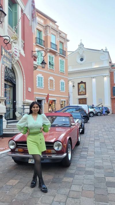 Traveler Jyoti beside a vintage red car in a pastel-coloured street of Monaco's old town, Monaco-Ville