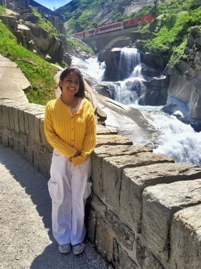 Traveler Jyoti standing by a waterfall in the Schöllenen Gorge with a red train crossing the bridge above near Andermatt
