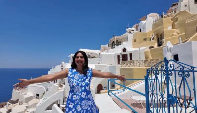 Traveler Jyoti on a clifftop terrace in Oia, Santorini, arms open, with white-and-blue Cycladic houses and the blue Aegean sea behind her