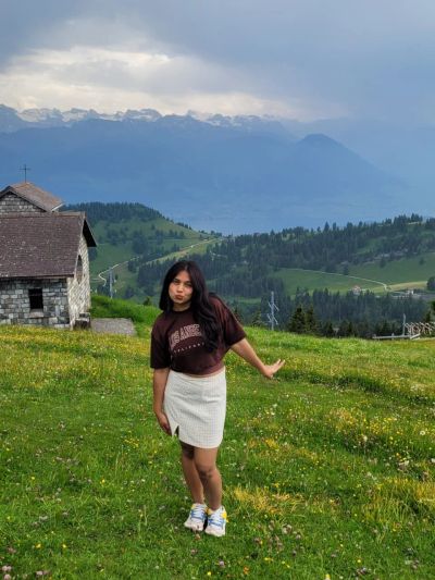 Traveler Jyoti on the green summit meadow of Mount Rigi with a small stone chapel and Lake Lucerne and the Alps behind her