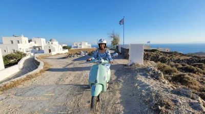 Traveler Jyoti riding a mint-green Vespa scooter on a Santorini clifftop path, with white Cycladic houses, a blue-domed church and the Greek flag behind her