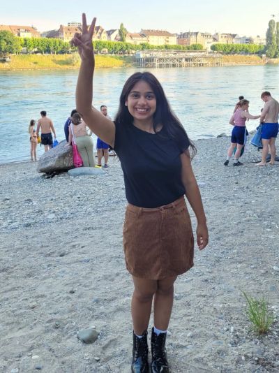 Traveler Jyoti on the pebbly bank of the Rhine River in Basel in summer, with locals swimming behind her