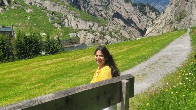 Traveler Jyoti seated on a wooden bench in the Schöllenen valley near Andermatt, with cliffs and green meadows behind
