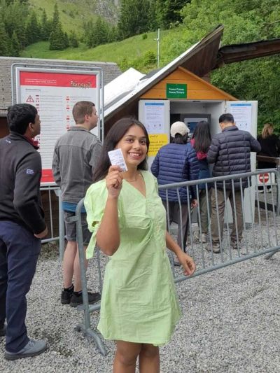 Traveler Jyoti holding a Fräkigaudi toboggan ticket in front of the self-service ticket machine at Fräkmüntegg