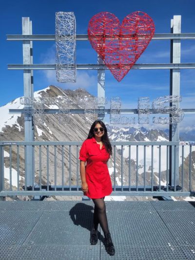 Traveler Jyoti in a red dress in front of the I love Titlis sign at the snowy Mount Titlis summit with blue sky